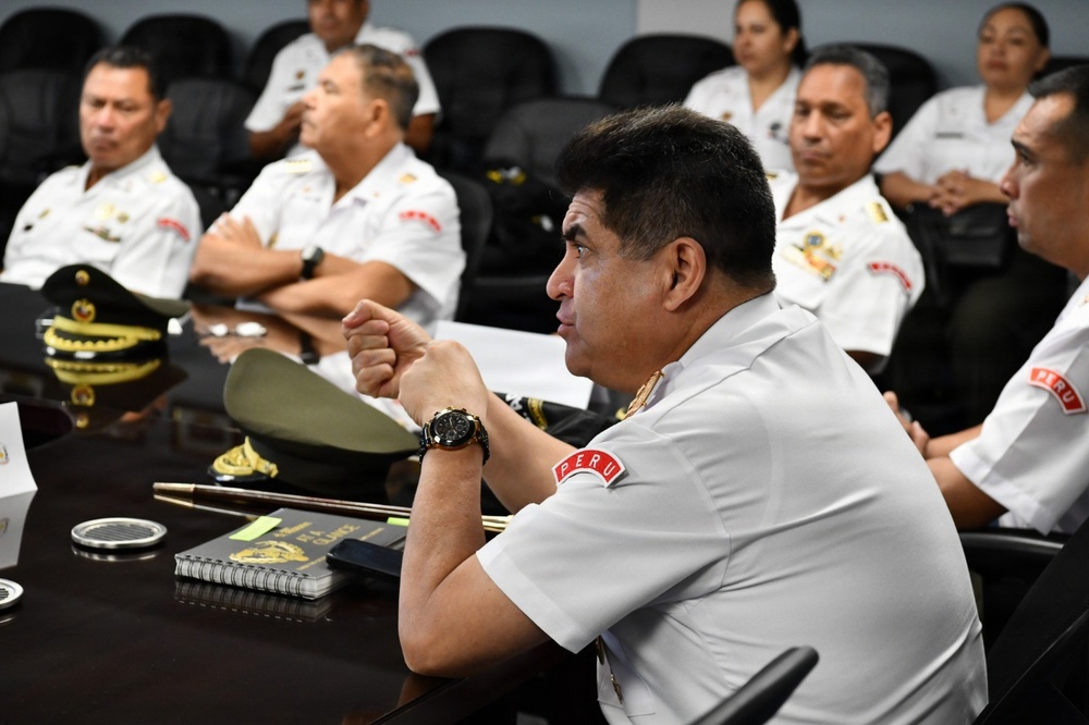 A group of Peruvian military or naval officers are seated in what appears to be a formal briefing or conference room. The personnel are wearing white uniforms with 'PERU' shoulder patches and various rank insignia. The setting suggests an official meeting, training session, or formal presentation.