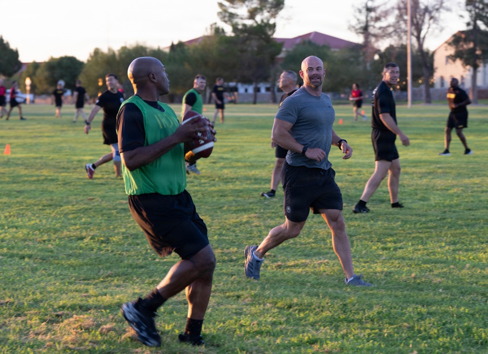 A group of adults are playing flag football on a grass field during golden hour lighting. Two teams are distinguishable, with one team wearing green mesh pinnies and the other in black shirts. The game appears to be an informal or recreational league, possibly on a military base given the surrounding buildings and field layout.