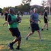 A group of adults are playing flag football on a grass field during golden hour lighting. Two teams are distinguishable, with one team wearing green mesh pinnies and the other in black shirts. The game appears to be an informal or recreational league, possibly on a military base given the surrounding buildings and field layout.