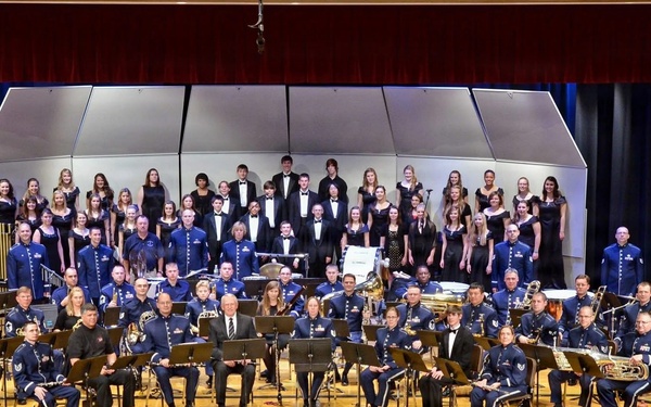A large formal group photograph taken on a concert hall stage featuring a military band in blue dress uniforms positioned in the foreground, with a civilian choir dressed in black formal attire arranged on risers behind them. The ensemble appears to be a U.S. Air Force band based on the distinctive blue uniforms, posing alongside what appears to be a school or community choir. The setting is a well-lit auditorium with red curtains, blue drapes, and a polished wooden stage floor.