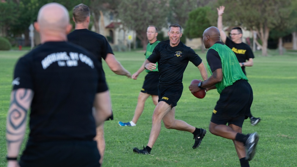 A group of individuals wearing U.S. Army-branded athletic clothing are playing flag football on a grass field. Two teams are distinguishable by their shirt colors — black and green. The game appears casual and recreational, taking place outdoors near trees and residential buildings.