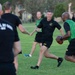 A group of individuals wearing U.S. Army-branded athletic clothing are playing flag football on a grass field. Two teams are distinguishable by their shirt colors — black and green. The game appears casual and recreational, taking place outdoors near trees and residential buildings.