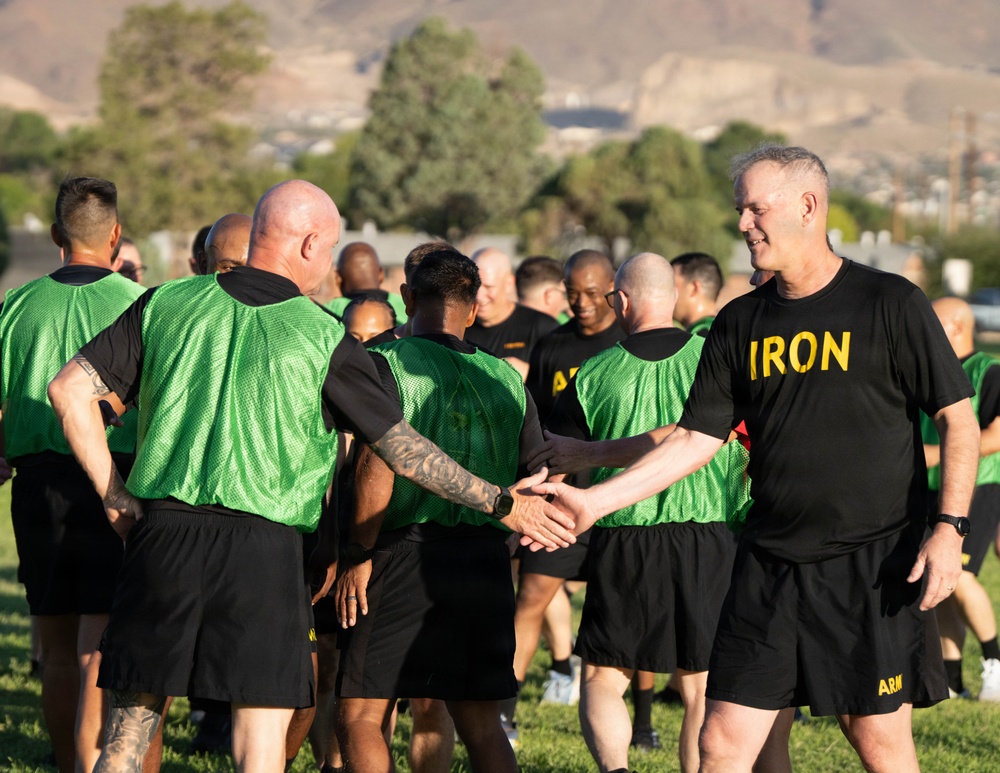 A group of military personnel in physical training attire are gathered on a grass field, likely during a unit PT (physical training) session. A senior individual wearing a black 'IRON' shirt is shaking hands with another soldier wearing a green mesh vest, suggesting a greeting or post-exercise interaction. The setting appears to be a military installation with arid hills visible in the background.
