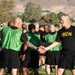 A group of military personnel in physical training attire are gathered on a grass field, likely during a unit PT (physical training) session. A senior individual wearing a black 'IRON' shirt is shaking hands with another soldier wearing a green mesh vest, suggesting a greeting or post-exercise interaction. The setting appears to be a military installation with arid hills visible in the background.