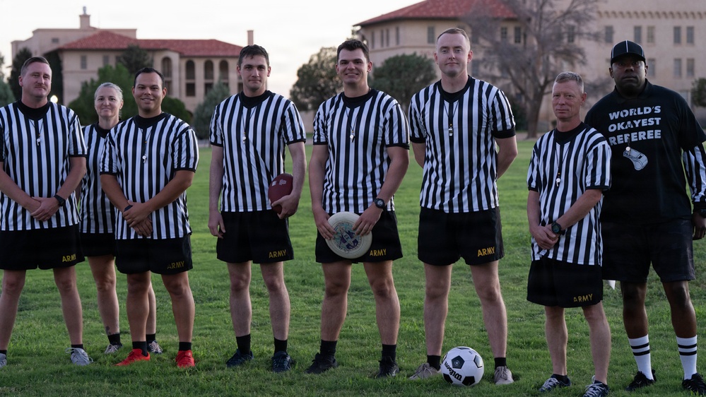 A group of eight individuals pose on a grass field wearing black-and-white striped referee shirts over black Army-branded shorts. Most appear to be U.S. Army personnel, with one person on the far right wearing a novelty shirt reading 'World's Okayest Referee.' Several sports items are visible including a soccer ball, a frisbee, and an American football, suggesting a multi-sport officiating event.