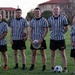 A group of eight individuals pose on a grass field wearing black-and-white striped referee shirts over black Army-branded shorts. Most appear to be U.S. Army personnel, with one person on the far right wearing a novelty shirt reading 'World's Okayest Referee.' Several sports items are visible including a soccer ball, a frisbee, and an American football, suggesting a multi-sport officiating event.