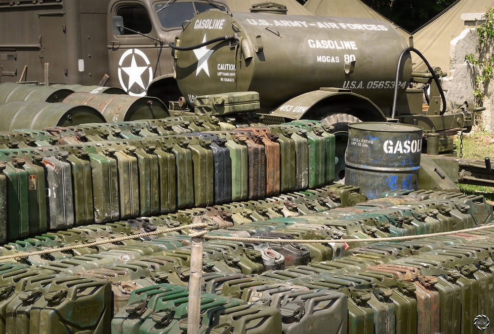 A large collection of military jerry cans arranged in rows on display, likely at a military history museum or reenactment event. Behind the cans are U.S. Army Air Forces gasoline tanker trucks marked with white stars and identification numbers. The scene depicts WWII-era American military fuel supply equipment.