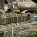 A large collection of military jerry cans arranged in rows on display, likely at a military history museum or reenactment event. Behind the cans are U.S. Army Air Forces gasoline tanker trucks marked with white stars and identification numbers. The scene depicts WWII-era American military fuel supply equipment.