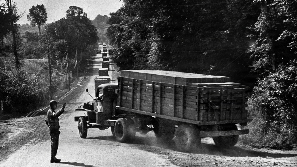 A black-and-white archival photograph showing a military supply convoy of cargo trucks traveling along a tree-lined road. A uniformed soldier stands at the roadside, appearing to direct or signal the convoy traffic. The trucks are loaded with stacked wooden crates or boxes, suggesting a military logistics operation.