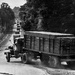 A black-and-white archival photograph showing a military supply convoy of cargo trucks traveling along a tree-lined road. A uniformed soldier stands at the roadside, appearing to direct or signal the convoy traffic. The trucks are loaded with stacked wooden crates or boxes, suggesting a military logistics operation.