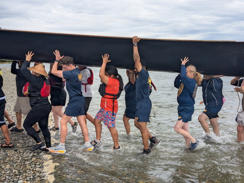 A group of people, several wearing navy blue uniforms with 'NAVY' branding, are collectively carrying a large black boat or canoe overhead toward the water's edge. The team is wading through shallow water on a rocky shoreline, working together in a coordinated effort to launch the vessel. The scene appears to be a military or team-building exercise involving watercraft handling.
