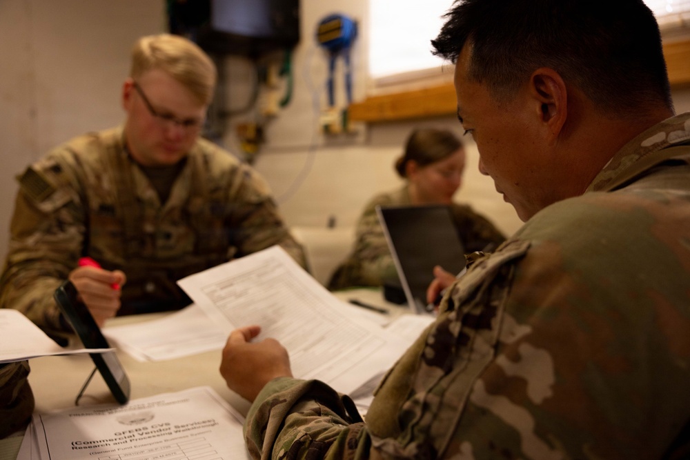 Three military personnel in camouflage uniforms are seated at a table engaged in administrative work. The foreground soldier is reviewing a document labeled 'GFEBS CVS (Commercial Vendor Services),' while others work with a tablet and laptop. The setting appears to be an indoor military workspace or operations room.