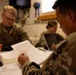 Three military personnel in camouflage uniforms are seated at a table engaged in administrative work. The foreground soldier is reviewing a document labeled 'GFEBS CVS (Commercial Vendor Services),' while others work with a tablet and laptop. The setting appears to be an indoor military workspace or operations room.