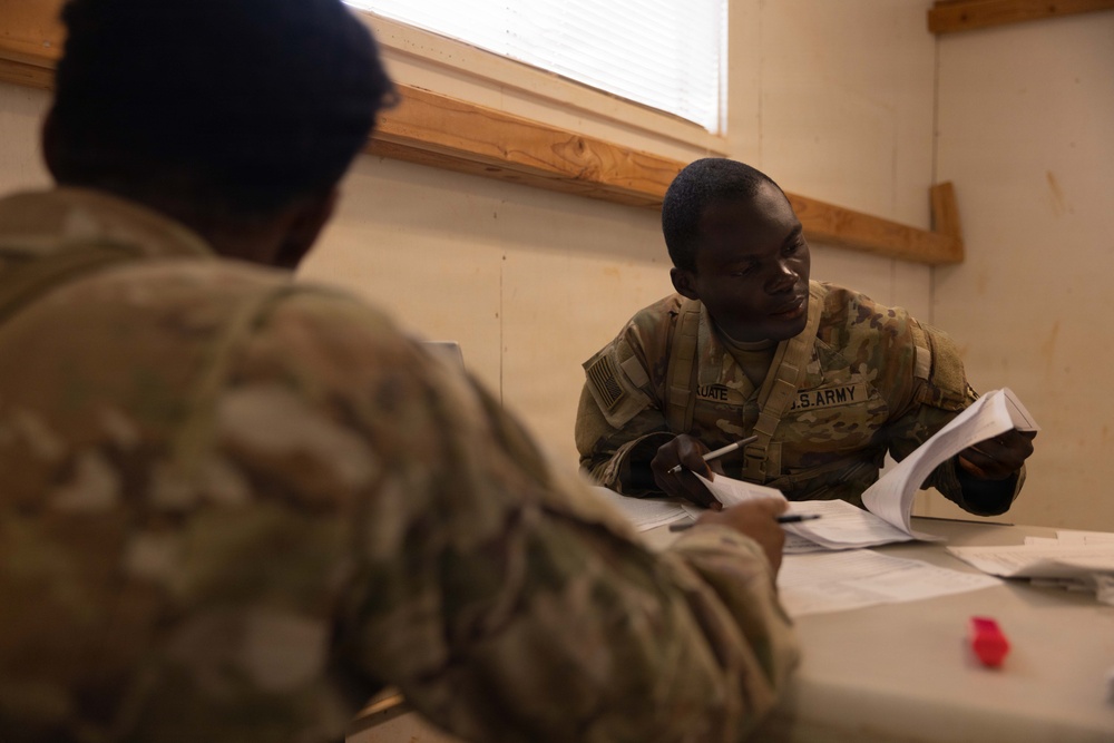 Two U.S. Army soldiers in OCP (Operational Camouflage Pattern) uniforms are seated at a table engaged in paperwork processing. The soldier facing the camera has a name tape partially readable as 'KUATE' and a 'U.S. ARMY' chest tape visible. The setting appears to be a temporary or forward operating base administrative area with plywood construction visible.