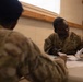 Two U.S. Army soldiers in OCP (Operational Camouflage Pattern) uniforms are seated at a table engaged in paperwork processing. The soldier facing the camera has a name tape partially readable as 'KUATE' and a 'U.S. ARMY' chest tape visible. The setting appears to be a temporary or forward operating base administrative area with plywood construction visible.