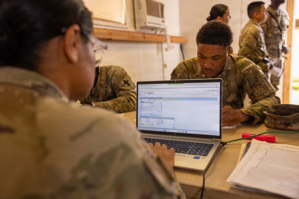 Two U.S. Army soldiers in OCP (Operational Camouflage Pattern) uniforms are working together at a table with an HP laptop displaying what appears to be a military logistics or administrative software interface. A second soldier leans in to observe the screen while the first operates the keyboard. Additional uniformed personnel are visible in the background of what appears to be a temporary or field operations building.