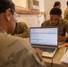 Two U.S. Army soldiers in OCP (Operational Camouflage Pattern) uniforms are working together at a table with an HP laptop displaying what appears to be a military logistics or administrative software interface. A second soldier leans in to observe the screen while the first operates the keyboard. Additional uniformed personnel are visible in the background of what appears to be a temporary or field operations building.