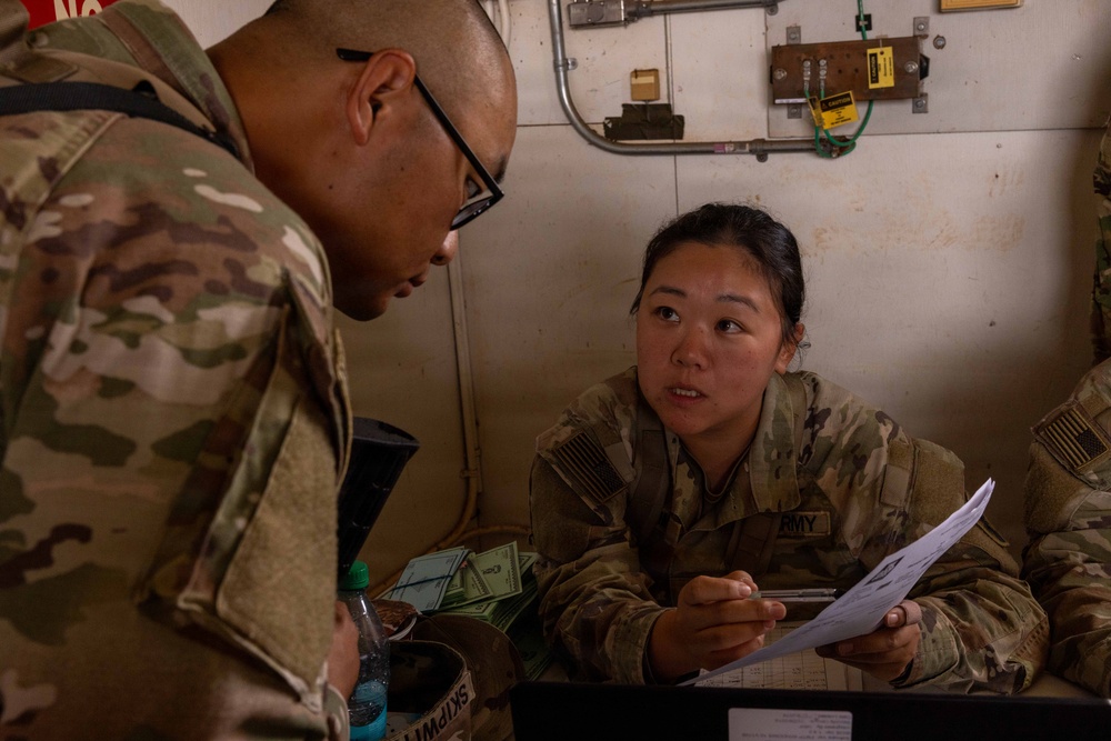 Two U.S. Army soldiers in MultiCam/OCP camouflage uniforms are engaged in what appears to be an administrative or finance transaction at a desk. A female soldier seated at the desk is holding a clipboard with documents and speaking with a male soldier wearing glasses who is leaning over the desk. Currency (U.S. dollars) is visible on the desk, suggesting a finance or pay operation.