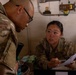 Two U.S. Army soldiers in MultiCam/OCP camouflage uniforms are engaged in what appears to be an administrative or finance transaction at a desk. A female soldier seated at the desk is holding a clipboard with documents and speaking with a male soldier wearing glasses who is leaning over the desk. Currency (U.S. dollars) is visible on the desk, suggesting a finance or pay operation.