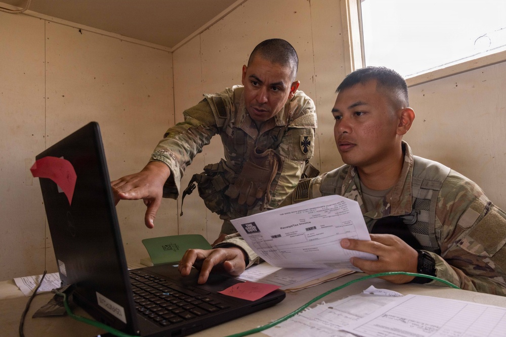 Two U.S. Army soldiers in MultiCam/OCP camouflage uniforms are working together at a desk in what appears to be a forward operating base or temporary military facility. One soldier is pointing at a laptop screen while the other holds printed documents, suggesting they are reviewing administrative or logistical paperwork. The setting appears to be an austere military workspace with minimal furnishings.
