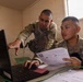 Two U.S. Army soldiers in MultiCam/OCP camouflage uniforms are working together at a desk in what appears to be a forward operating base or temporary military facility. One soldier is pointing at a laptop screen while the other holds printed documents, suggesting they are reviewing administrative or logistical paperwork. The setting appears to be an austere military workspace with minimal furnishings.