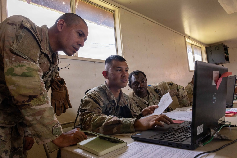 Three military personnel in U.S. Army OCP (Operational Camouflage Pattern) uniforms are gathered around a desk reviewing information on a Dell laptop computer. One soldier is seated at the workstation while the other two lean in to observe the screen, with one holding a document. The setting appears to be a forward operating base or temporary command post interior.