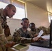 Three military personnel in U.S. Army OCP (Operational Camouflage Pattern) uniforms are gathered around a desk reviewing information on a Dell laptop computer. One soldier is seated at the workstation while the other two lean in to observe the screen, with one holding a document. The setting appears to be a forward operating base or temporary command post interior.