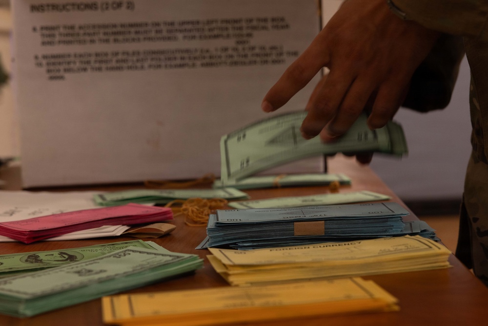 A person in military/camouflage uniform is handling what appears to be currency and documents on a wooden table. Multiple stacks of bills, envelopes, and paperwork are spread across the surface, with a visible 'FOREIGN CURRENCY' labeled envelope and an instruction sheet in the background. The scene suggests an official currency counting, processing, or evidence documentation activity.