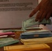 A person in military/camouflage uniform is handling what appears to be currency and documents on a wooden table. Multiple stacks of bills, envelopes, and paperwork are spread across the surface, with a visible 'FOREIGN CURRENCY' labeled envelope and an instruction sheet in the background. The scene suggests an official currency counting, processing, or evidence documentation activity.