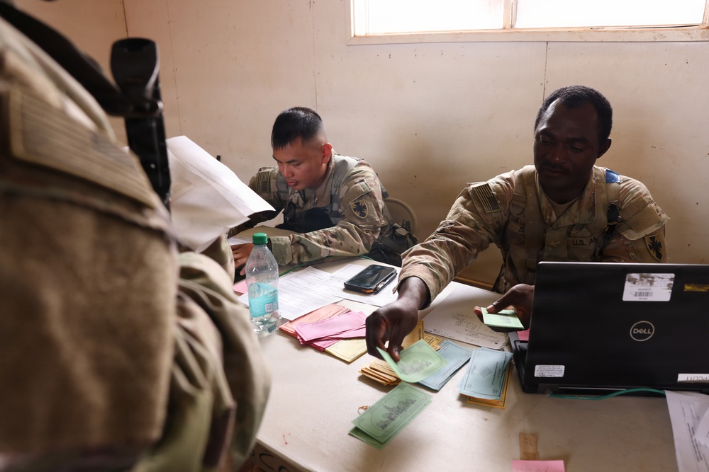 Two U.S. Army soldiers in camouflage uniforms are seated at a desk covered with documents, colored papers, and a Dell laptop, appearing to process paperwork or conduct an administrative interview. A third individual, seen from behind in the foreground, is interacting with the soldiers across the desk. The setting appears to be a temporary or forward operating base office environment.