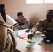 Two U.S. Army soldiers in camouflage uniforms are seated at a desk covered with documents, colored papers, and a Dell laptop, appearing to process paperwork or conduct an administrative interview. A third individual, seen from behind in the foreground, is interacting with the soldiers across the desk. The setting appears to be a temporary or forward operating base office environment.