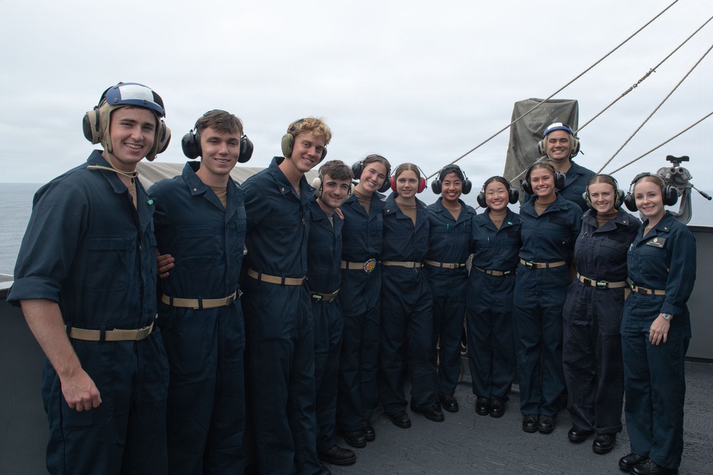 A group of approximately 11 young naval personnel in dark blue coveralls pose together on the deck of a military vessel at sea. Most individuals are wearing hearing protection/headsets, suggesting they are or were recently in a high-noise environment such as a flight deck. The group appears to be in good spirits, smiling for a group photograph.
