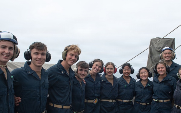 A group of approximately 11 young naval personnel in dark blue coveralls pose together on the deck of a military vessel at sea. Most individuals are wearing hearing protection/headsets, suggesting they are or were recently in a high-noise environment such as a flight deck. The group appears to be in good spirits, smiling for a group photograph.