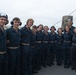 A group of approximately 11 young naval personnel in dark blue coveralls pose together on the deck of a military vessel at sea. Most individuals are wearing hearing protection/headsets, suggesting they are or were recently in a high-noise environment such as a flight deck. The group appears to be in good spirits, smiling for a group photograph.