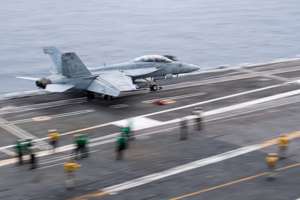 A U.S. Navy fighter jet, appearing to be an F/A-18 Hornet or Super Hornet, is captured mid-launch or landing on an aircraft carrier flight deck. The image shows significant motion blur, indicating high speed during the operation. Deck crew members in colored jerseys are visible in the foreground, consistent with standard carrier flight deck operations.