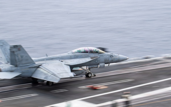 A U.S. Navy fighter jet, appearing to be an F/A-18 Hornet or Super Hornet, is captured mid-launch or landing on an aircraft carrier flight deck. The image shows significant motion blur, indicating high speed during the operation. Deck crew members in colored jerseys are visible in the foreground, consistent with standard carrier flight deck operations.