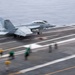 A U.S. Navy fighter jet, appearing to be an F/A-18 Hornet or Super Hornet, is captured mid-launch or landing on an aircraft carrier flight deck. The image shows significant motion blur, indicating high speed during the operation. Deck crew members in colored jerseys are visible in the foreground, consistent with standard carrier flight deck operations.