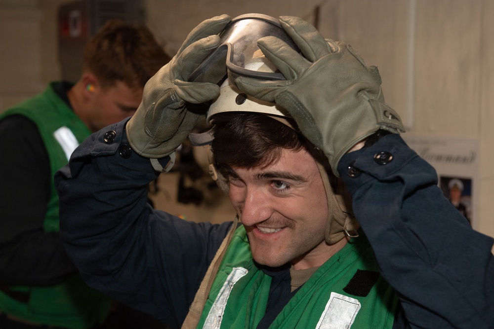 A young man wearing a green safety vest and olive-colored work gloves is placing or adjusting a helmet with a visor on his head while smiling. He appears to be in an indoor industrial or military setting. A second person in a green vest is visible in the background, slightly out of focus.