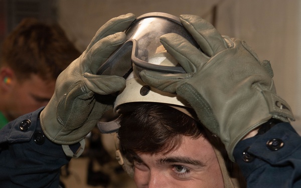 A young man wearing a green safety vest and olive-colored work gloves is placing or adjusting a helmet with a visor on his head while smiling. He appears to be in an indoor industrial or military setting. A second person in a green vest is visible in the background, slightly out of focus.