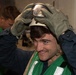 A young man wearing a green safety vest and olive-colored work gloves is placing or adjusting a helmet with a visor on his head while smiling. He appears to be in an indoor industrial or military setting. A second person in a green vest is visible in the background, slightly out of focus.