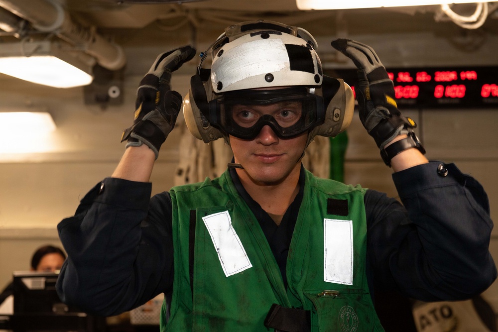 A military personnel member, likely a U.S. Navy sailor, is shown wearing flight deck safety gear including a white helmet, ear protection/headset, protective goggles, and a green safety vest. The individual is adjusting their helmet with both gloved hands while standing in what appears to be an interior compartment of a naval vessel. A second person is partially visible in the background near a workstation.