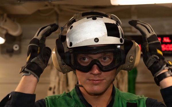 A military personnel member, likely a U.S. Navy sailor, is shown wearing flight deck safety gear including a white helmet, ear protection/headset, protective goggles, and a green safety vest. The individual is adjusting their helmet with both gloved hands while standing in what appears to be an interior compartment of a naval vessel. A second person is partially visible in the background near a workstation.