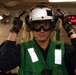 A military personnel member, likely a U.S. Navy sailor, is shown wearing flight deck safety gear including a white helmet, ear protection/headset, protective goggles, and a green safety vest. The individual is adjusting their helmet with both gloved hands while standing in what appears to be an interior compartment of a naval vessel. A second person is partially visible in the background near a workstation.