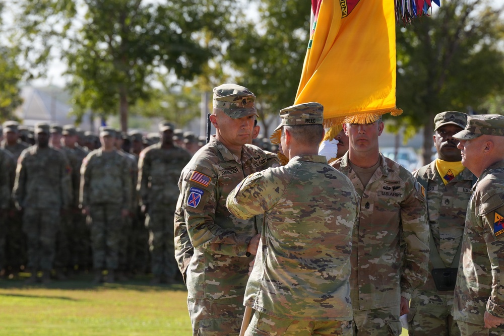 A U.S. Army military ceremony, likely a change of command or activation ceremony, is taking place outdoors on a parade field. Soldiers in Army Combat Uniforms (ACUs/OCP pattern) are passing or receiving a unit guidon (yellow flag), which is the central symbolic act of such ceremonies. Formations of soldiers stand at attention in the background.