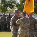 A U.S. Army military ceremony, likely a change of command or activation ceremony, is taking place outdoors on a parade field. Soldiers in Army Combat Uniforms (ACUs/OCP pattern) are passing or receiving a unit guidon (yellow flag), which is the central symbolic act of such ceremonies. Formations of soldiers stand at attention in the background.