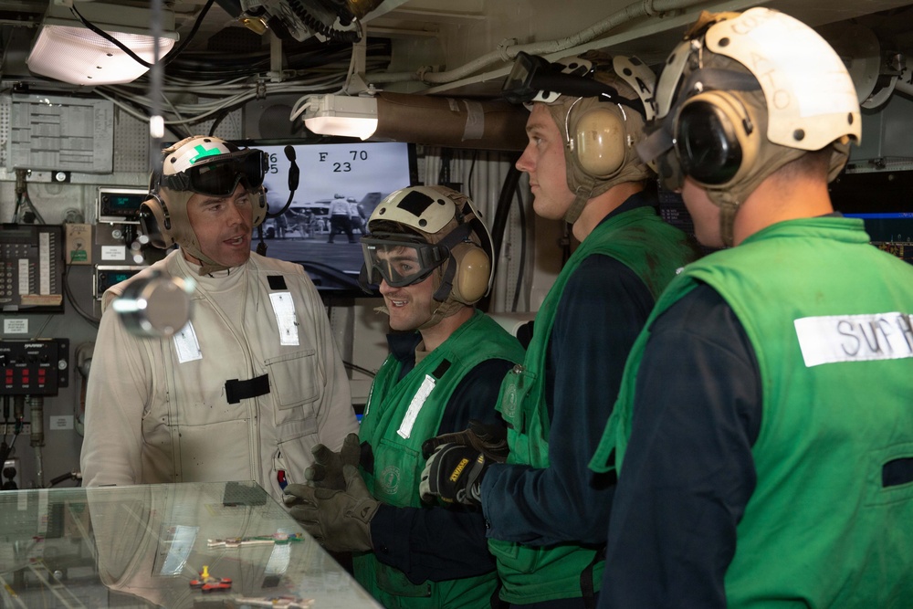 The image shows military personnel, likely U.S. Navy sailors, gathered in what appears to be a ship's flight deck control room or similar operational space aboard an aircraft carrier. One individual in a tan/white flight suit is briefing or conversing with three others wearing green flight deck jerseys and helmets with hearing protection. A monitor in the background displays a black-and-white feed of flight deck operations.