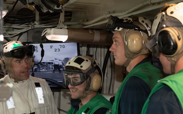 The image shows military personnel, likely U.S. Navy sailors, gathered in what appears to be a ship's flight deck control room or similar operational space aboard an aircraft carrier. One individual in a tan/white flight suit is briefing or conversing with three others wearing green flight deck jerseys and helmets with hearing protection. A monitor in the background displays a black-and-white feed of flight deck operations.