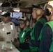 The image shows military personnel, likely U.S. Navy sailors, gathered in what appears to be a ship's flight deck control room or similar operational space aboard an aircraft carrier. One individual in a tan/white flight suit is briefing or conversing with three others wearing green flight deck jerseys and helmets with hearing protection. A monitor in the background displays a black-and-white feed of flight deck operations.