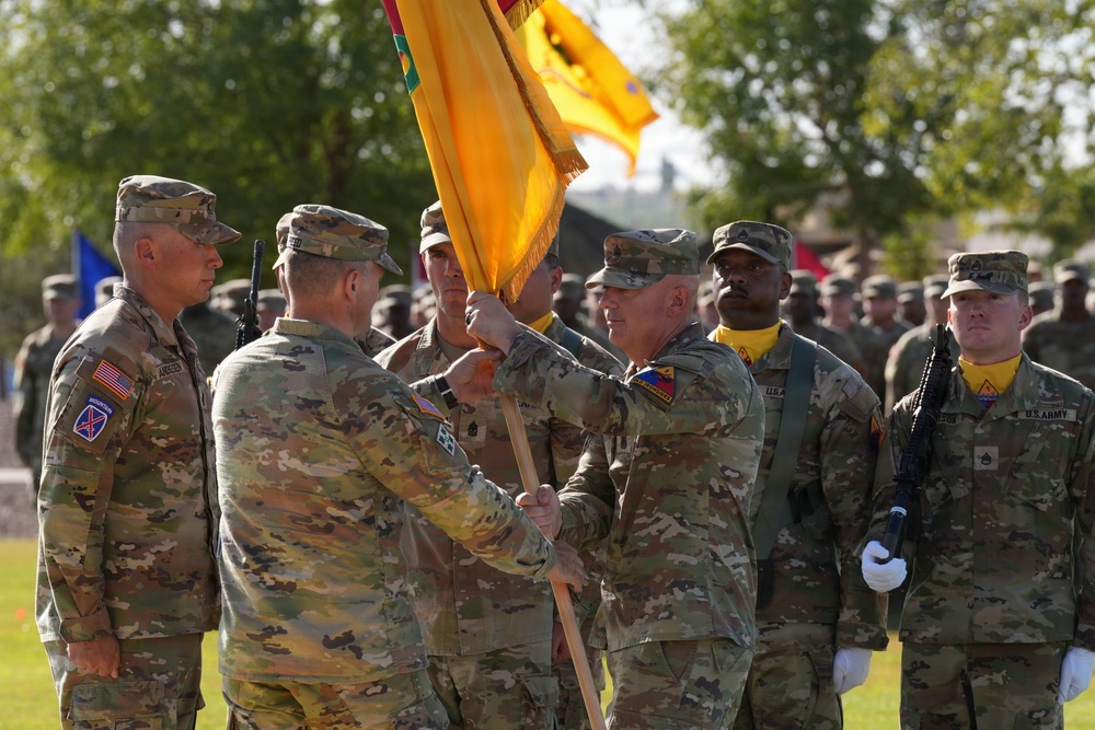 A U.S. Army change of command ceremony is taking place outdoors, with officers passing a unit guidon (flag) between them, symbolizing the transfer of command authority. Soldiers in Army Combat Uniforms (ACU/OCP pattern) are assembled in formation in the background. The ceremony appears to be conducted on a parade field with trees visible in the background.