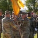 A U.S. Army change of command ceremony is taking place outdoors, with officers passing a unit guidon (flag) between them, symbolizing the transfer of command authority. Soldiers in Army Combat Uniforms (ACU/OCP pattern) are assembled in formation in the background. The ceremony appears to be conducted on a parade field with trees visible in the background.